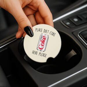 May include: A round, off-white car coaster with the words "PLACE DIET COKE HERE PLEASE" printed around a graphic of a Diet Coke can. The coaster is being held in a car's cup holder.