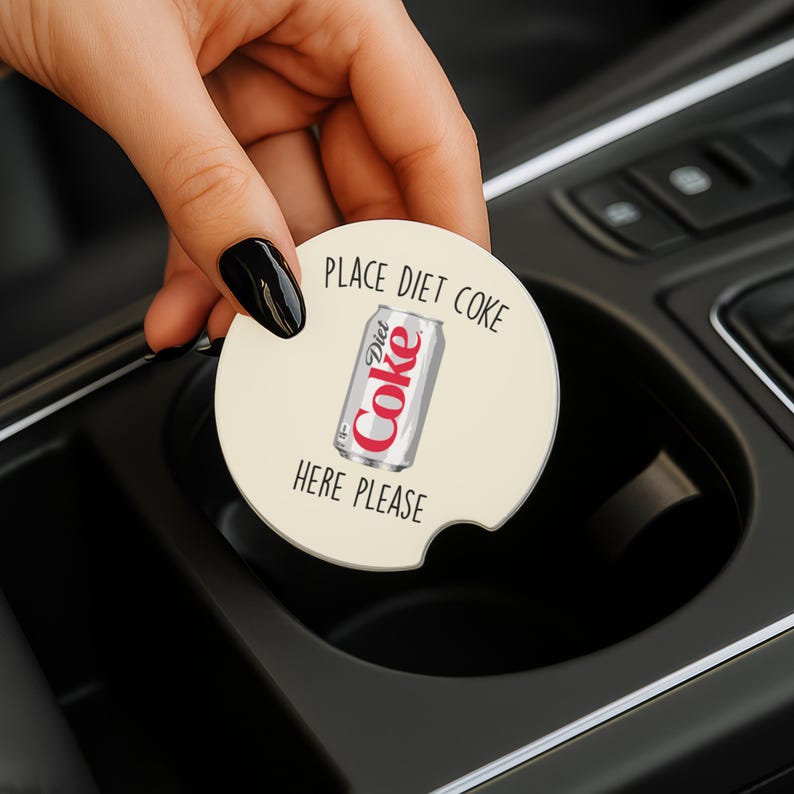 May include: A round, off-white car coaster with the words "PLACE DIET COKE HERE PLEASE" printed around a graphic of a Diet Coke can. The coaster is being held in a car's cup holder.