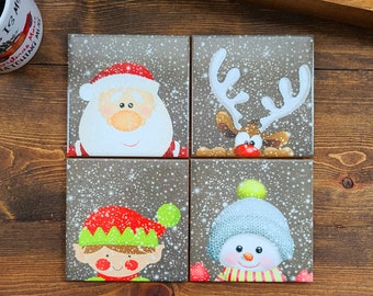 May include: Set of four square ceramic coasters featuring a winter holiday theme. Each coaster has a different character: Santa Claus, a reindeer, an elf, and a snowman. The coasters have a brown background with a white snowflake pattern.