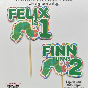 May include: Two colorful layered card cake toppers featuring the Very Hungry Caterpillar. The first topper says "Felix is 1" and the second topper says "Finn turns 2".