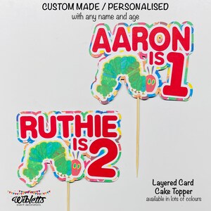 May include: Two colorful layered card cake toppers featuring the Very Hungry Caterpillar. The first topper says "AARON is 1" and the second topper says "RUTHIE is 2".