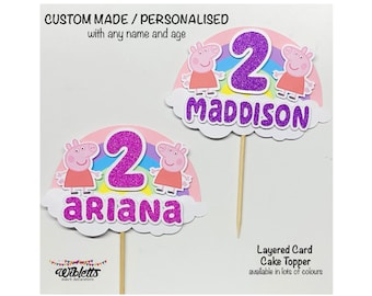 May include: Two pink pig character cake toppers with rainbow accents. The toppers are personalized with the names "Ariana" and "Maddison" and the number "2" in purple glitter. The toppers are on wooden skewers.