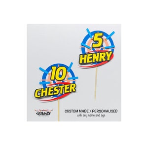 May include: Two personalised cake toppers with a target design. The top topper has a blue and red target with the number 5 and the name "HENRY" in black. The bottom topper has a blue and red target with the number 10 and the name "CHESTER" in black.