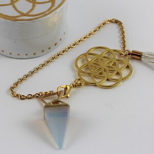 Puede incluir: Una caja redonda blanca con un diseño geométrico dorado en la tapa y puntos dorados alrededor de la base. Una cadena dorada con un péndulo y un dije dorado con una borla blanca están junto a la caja.