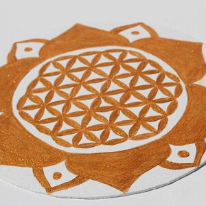 Könnte beinhalten: Ein goldfarbenes Mandala mit einem Lebensblume-Design, das auf eine weiße, runde Oberfläche gemalt ist.