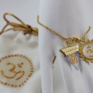 Könnte beinhalten: Ein goldfarbenes Kettenarmband mit zwei Anhängern: ein Sonnenstrahl und ein Dreieck, beide mit dem Om-Symbol. Das Armband wird auf einer weißen Hand präsentiert. Ein weißer Kordelzugbeutel mit dem Om-Symbol ist im Hintergrund.