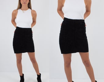 May include: A black suede mini skirt with a high waist and a straight silhouette. The skirt is shown on a model wearing a white tank top and black ankle boots.
