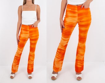 May include: A woman is wearing a white tank top and orange flared pants with a tie-dye pattern. She is wearing white sandals.