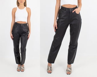 May include: A woman is wearing black leather pants with a high waist and a white tank top. She is wearing snakeskin sandals.
