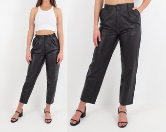 May include: Black leather trousers with a high waist and button fastening. The straight-leg trousers are worn with a white ribbed vest top and black sandals. The model is posing against a white backdrop.