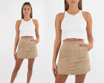 May include: A woman is wearing a white tank top and a light brown suede mini skirt with pockets. The skirt has a raw hem.