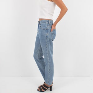 May include: A woman wearing a white tank top and light blue denim jeans with a straight leg fit. She is wearing black heeled sandals.