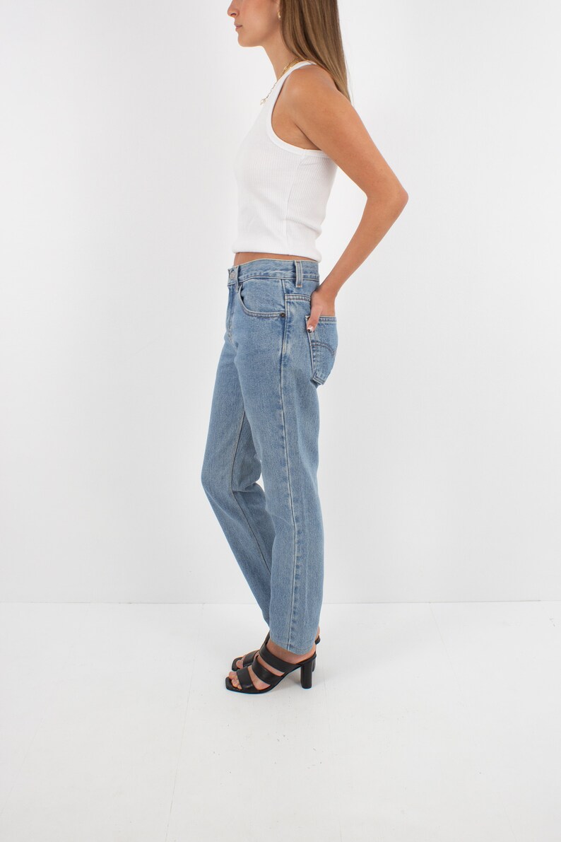 May include: A woman wearing a white tank top and light blue denim jeans with a straight leg fit. She is wearing black heeled sandals.