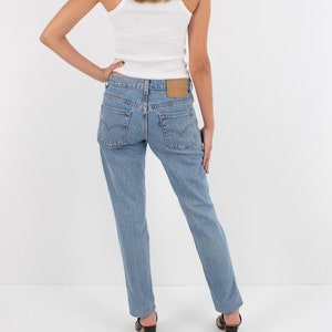 May include: A woman wearing a white tank top and light blue denim jeans. The jeans have a brown leather patch on the back with the Levi's logo.