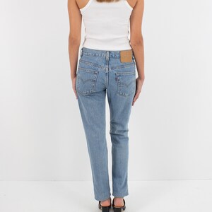 May include: A woman wearing a white tank top and light blue denim jeans with a brown leather patch on the back pocket. She is wearing black heeled mules.