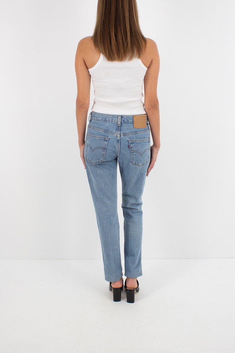 May include: A woman wearing a white tank top and light blue denim jeans with a brown leather patch on the back pocket. She is wearing black heeled mules.