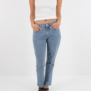 May include: A woman wearing a white ribbed tank top and light blue denim jeans. She is wearing black heeled sandals.