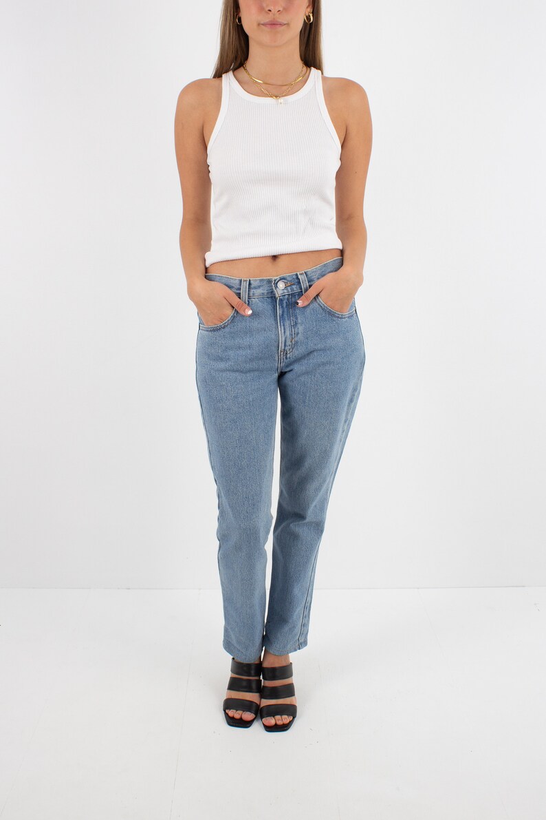 May include: A woman wearing a white ribbed tank top and light blue denim jeans. She is wearing black heeled sandals.