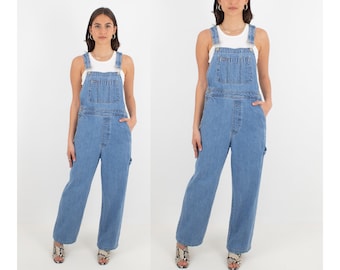 May include: Two women wearing light blue denim overalls with wide legs. Both women are wearing white tank tops and have their hands in their pockets.