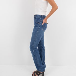 May include: A woman wearing a white tank top and blue denim jeans. She is standing with her hand in her pocket and wearing black heeled sandals.
