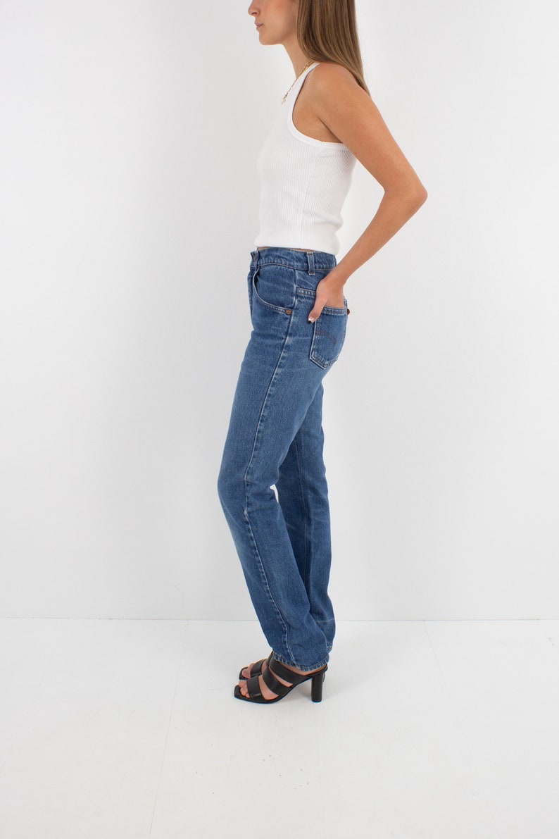 May include: A woman wearing a white tank top and blue denim jeans. She is standing with her hand in her pocket and wearing black heeled sandals.