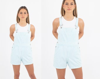 May include: Two women wearing light blue gingham overall shorts. The shorts have adjustable straps and are a casual style.