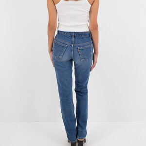 May include: A woman wearing a white tank top and blue denim jeans. The jeans have a high waist and a straight leg.