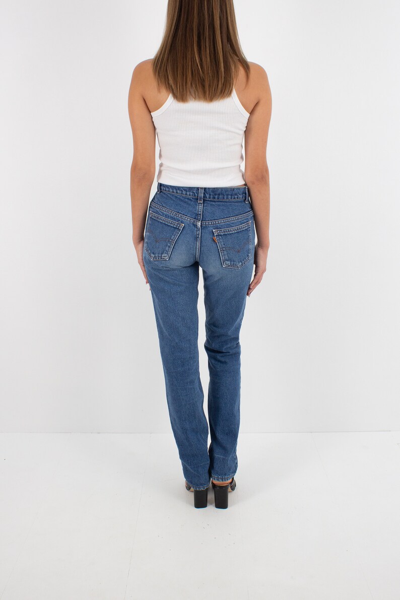 May include: A woman wearing a white tank top and blue denim jeans. The jeans have a high waist and a straight leg.
