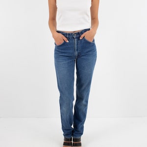 May include: A woman wearing a white tank top and blue denim jeans with a high waist. She is wearing black heeled sandals.