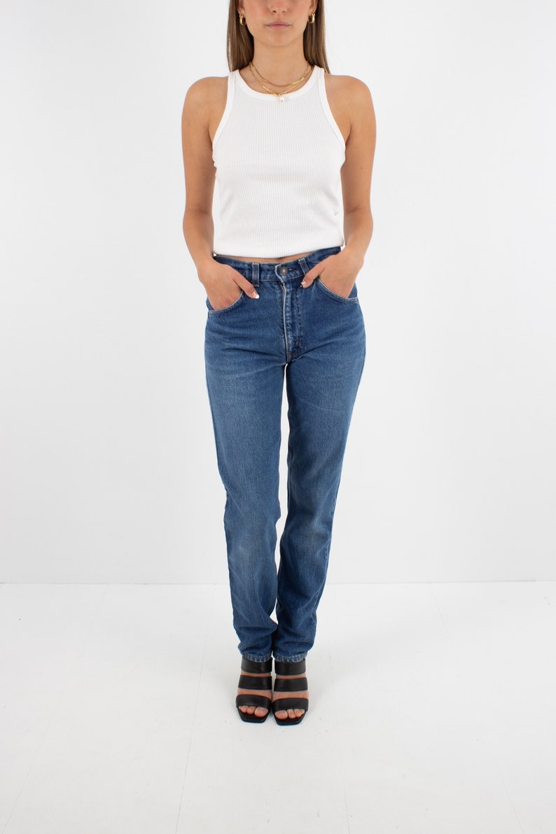 May include: A woman wearing a white tank top and blue denim jeans with a high waist. She is wearing black heeled sandals.