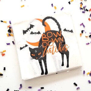 Puede incluir: Servilleta de papel blanca con una silueta de gato negro, una luna creciente y murciélagos. El texto "Bow Down Witches" está impreso en naranja y negro.