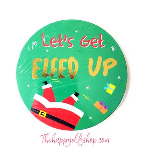 May include: A green round Christmas ornament with a white background and the text "Let's Get Elfed Up" in gold letters. A cartoon image of Santa Claus falling backwards is in the center of the ornament.