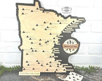 May include: A wooden map of Minnesota with green triangle markers indicating the location of state parks. The map is mounted on a black background with the text "Minnesota State Parks" in a circle. The map is surrounded by a white brick wall.