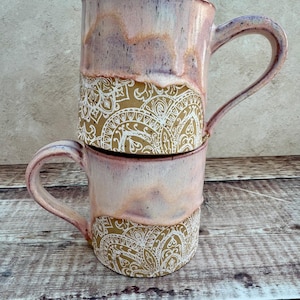 May include: Two stacked ceramic mugs. The mugs have a pink and peach glaze on the top half, and a tan base with a white paisley design. Each mug has a curved handle. The mugs are on a wooden surface.