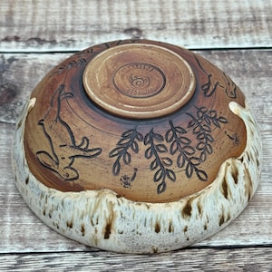 May include: A ceramic bowl with a brown, unglazed upper section featuring etched illustrations of a rabbit and leafy branches. The base is stamped with the text SeaBreeze Ceramics. The lower portion has a thick, textured white glaze drip.