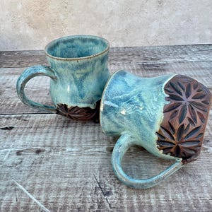 May include: Two ceramic mugs with a turquoise glaze and brown floral carved details. The mugs have curved handles and sit on a wooden surface. The mugs are handmade and suitable for coffee or tea.