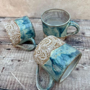 Peut inclure: Trois tasses en céramique présentant un design bicolore avec une finition émaillée bleue et blanche lisse sur la partie inférieure et une bande beige texturée sur le dessus ornée de motifs floraux blancs en relief et une anse courbée.