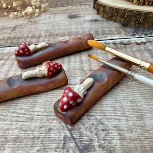 Peut inclure: Trois porte-pinceaux en céramique en forme de champignons avec des pois rouges et blancs. Les supports sont sur une surface en bois avec deux pinceaux.