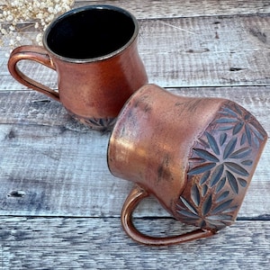 May include: Two copper-coloured ceramic mugs with handles. One mug is upright, the other is tilted, revealing a carved floral design on the side. The interior of the mugs is black. The mugs have a rustic, handmade appearance.
