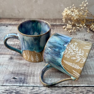 May include: Two handmade ceramic mugs. The mugs have a blue and teal glaze on the top half and a tan base. White floral designs decorate the base of one mug. The mugs have curved handles.