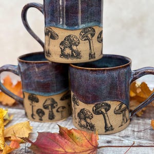 May include: Three ceramic mugs stacked, each with a dark blue glazed top and a tan base. The base features a black and white mushroom illustration. The mugs have large handles and are surrounded by autumn leaves.