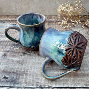 May include: Two handmade ceramic mugs with a blue and green glaze. One mug features a carved floral design in brown, while the other has a smooth exterior. Both mugs have curved handles and are set on a wooden surface.
