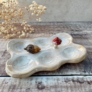 May include: A ceramic, off-white, cloud-shaped dish with six compartments. It features a small, red-capped mushroom and a brown acorn as decorative elements. The dish is placed on a wooden surface, with dried flowers in the background.