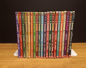 May include: A selection of Goosebumps books by R.L. Stine, with vibrant spines displaying titles such as "Stay Out of the Basement" and "Say Cheese and Die!" The books are displayed on a white book stand against a dark backdrop.