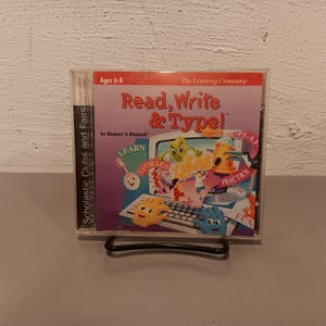 May include: A CD case for the computer game "Read, Write & Type!" for Windows and Macintosh. The case is pink and blue with cartoon characters and the text "The Learning Company" and "Scholastic Clubs and Fairs".