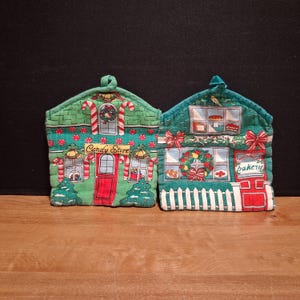 May include: Two house-shaped fabric pot holders featuring festive holiday designs. The left one is green with red accents, labeled Candy Store, and decorated with candy canes. The right one is green with red accents, labeled bakery, and features a wreath.