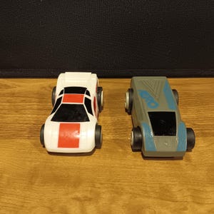May include: Two toy cars. One is white with a red stripe and black windows. The other is gray with blue accents and the word "Aero" printed on top. Both have black wheels and are on a wooden surface.