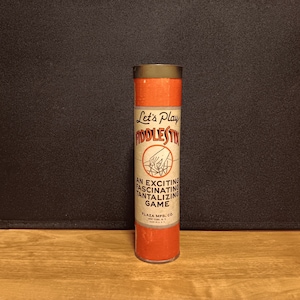 May include: A vintage cylindrical game container for "Fiddle Stix." The container is primarily orange and tan with a dark metal cap. The label reads "Let's Play Fiddle Stix" with additional text: "An exciting fascinating tantalizing game."