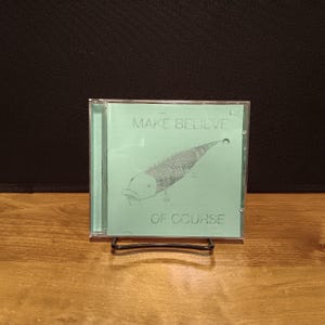 May include: A clear plastic CD jewel case featuring a light green cover insert. The text "MAKE BELIEVE" is printed at the top and "OF COURSE" at the bottom. A central illustration depicts a fish with a scaly, lizard-like tail and small legs.