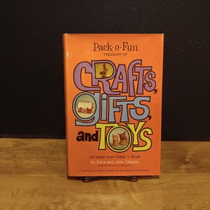 May include: An orange book titled "Pack-o-Fun Treasury of Crafts, Gifts and Toys." The cover features colorful text and illustrations of crafts. The book is made from odds and ends and is by Edna and John Clapper.